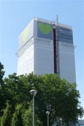 Uma imagem da Grenfell Tower após o incêndio de 2017, com um outdoor branco e verde erguido ao redor dela com os dizeres 