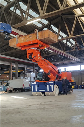 Guindaste industrial elétrico Ormig 100iE de alta resistência, na cor laranja, com a lança recolhida e a plataforma giratória carregada, dentro de uma fábrica.