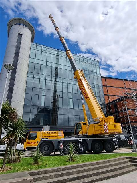 Side view of yellow five axle all terrain crane, boom up in front of a big glass building