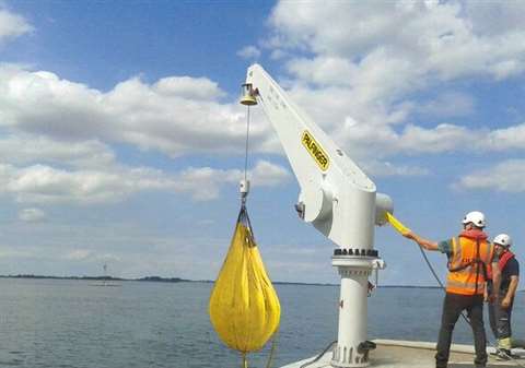 White fixed boom Palfinger crane installed by the sea with what looks like a yellow water weights test bag hanging from the hook and a couple of dudes in attendance